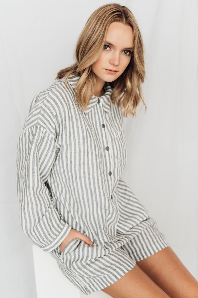 May include: A woman wearing a white and gray striped linen shirt dress with a button-up front and a pocket on the left side.