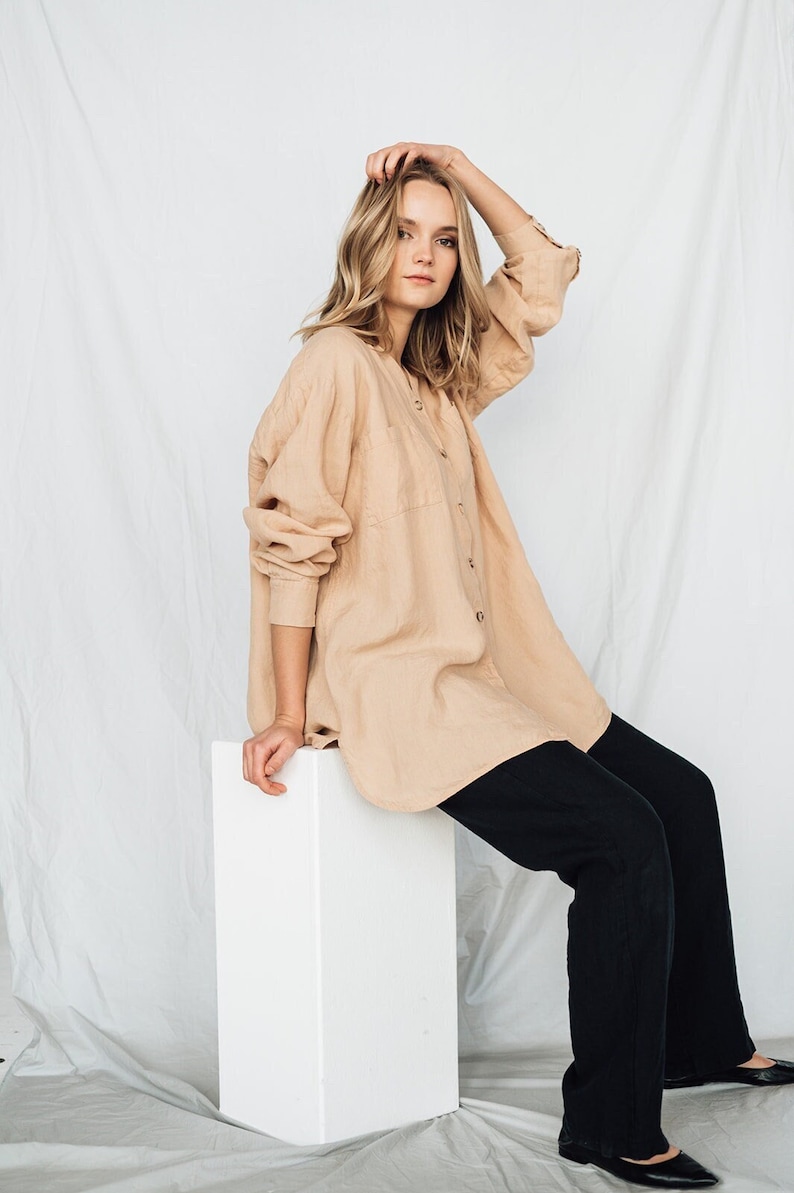 May include: A beige button-down shirt with long sleeves and a chest pocket, paired with black pants. The shirt is worn over the pants. The model is sitting on a white block.