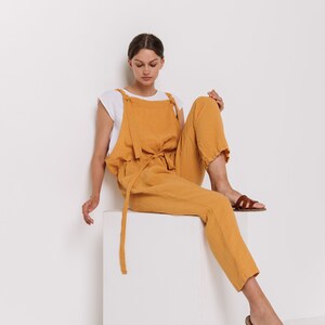 May include: A woman wearing a mustard yellow linen jumpsuit with a white undershirt and brown sandals sits on a white cube.