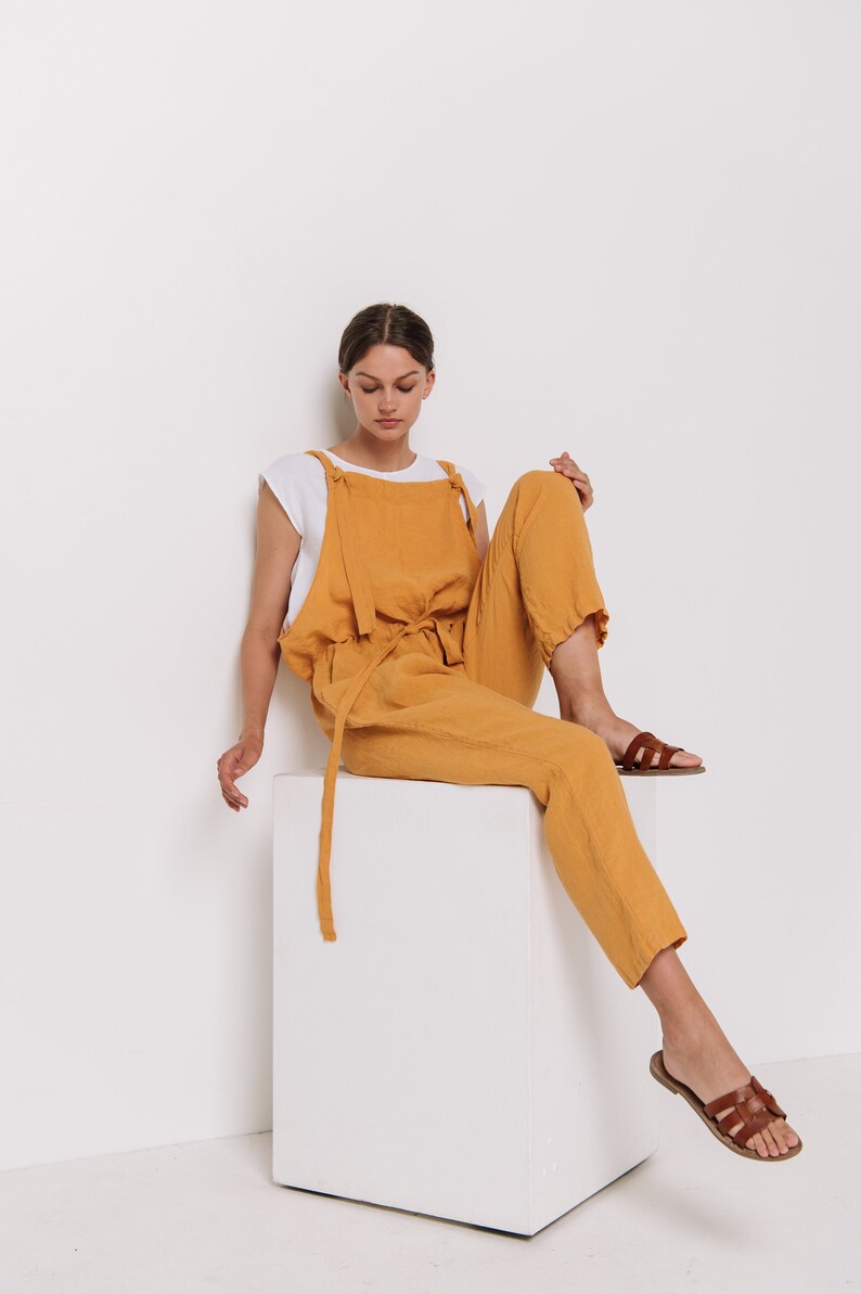May include: A woman wearing a mustard yellow linen jumpsuit with a white undershirt and brown sandals sits on a white cube.