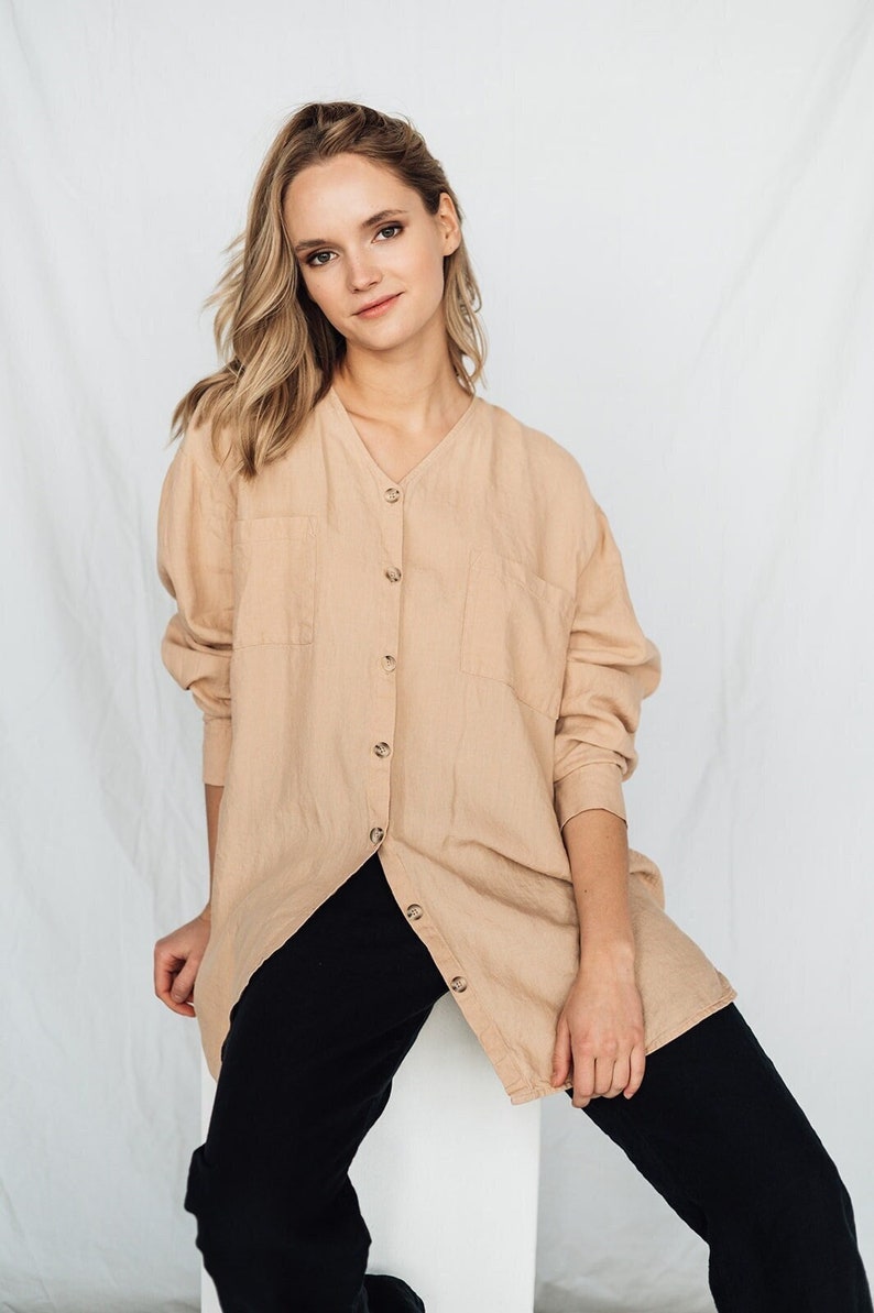 May include: A beige linen shirt with a V-neck and button-down front, paired with black pants. The shirt has two front pockets and long sleeves. The model is sitting on a white block against a white backdrop.