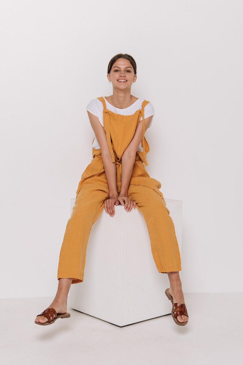 May include: A woman wearing a white t-shirt and mustard yellow overalls sits on a white cube. She is wearing brown sandals.
