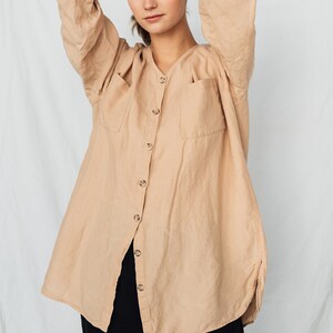 May include: A tan linen button-down shirt with two front pockets and long sleeves. The shirt is paired with black pants. The model is wearing the shirt with their arms behind their head.