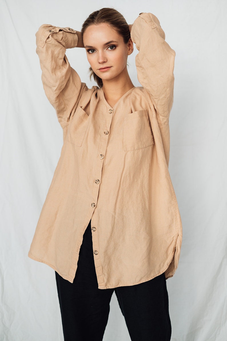 May include: A tan linen button-down shirt with two front pockets and long sleeves. The shirt is paired with black pants. The model is wearing the shirt with their arms behind their head.