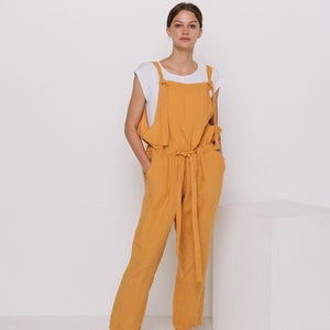 May include: A woman wearing a mustard yellow linen jumpsuit with a white tank top underneath. The jumpsuit has a tie at the waist and wide legs. She is wearing brown leather sandals.