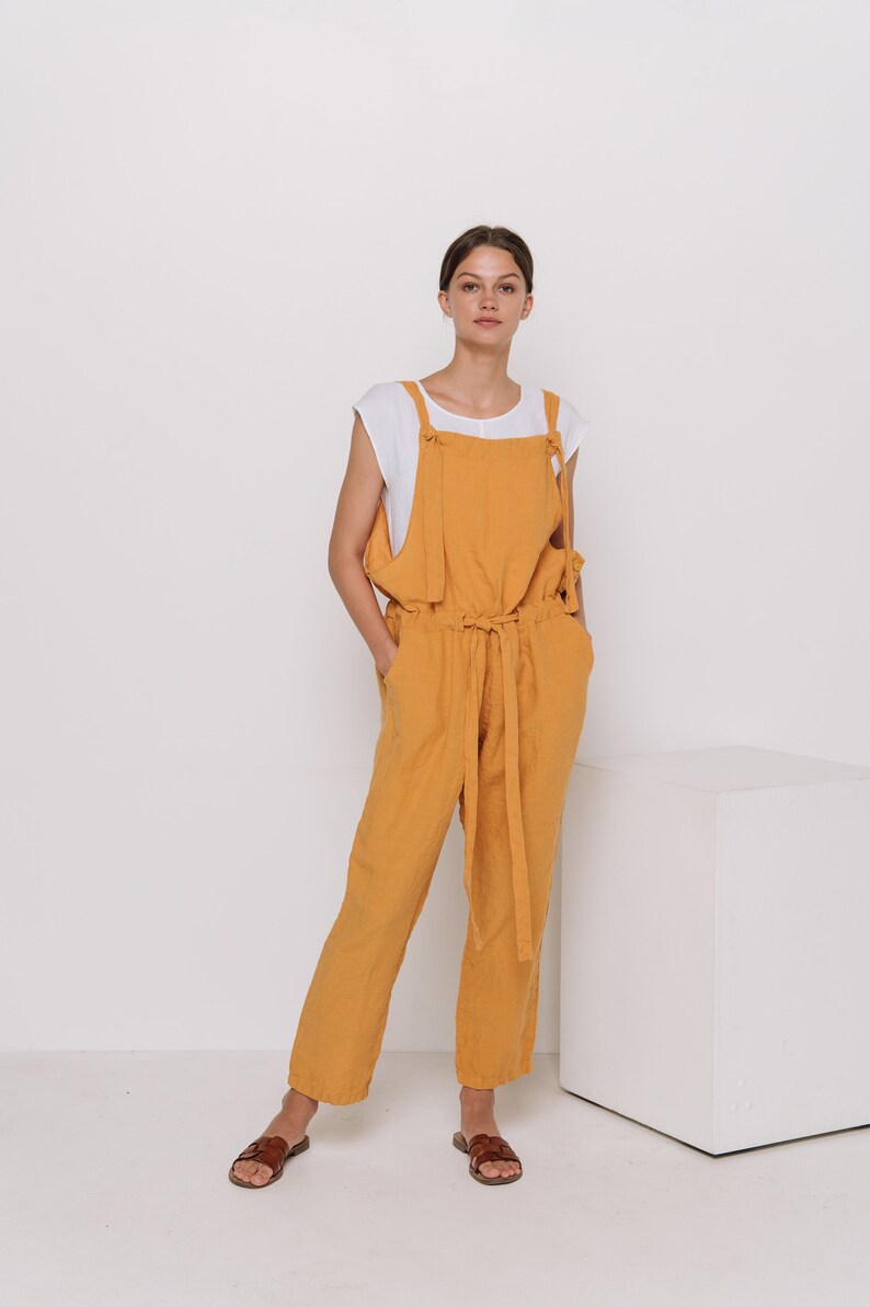 May include: A woman wearing a mustard yellow linen jumpsuit with a white tank top underneath. The jumpsuit has a tie at the waist and wide legs. She is wearing brown leather sandals.