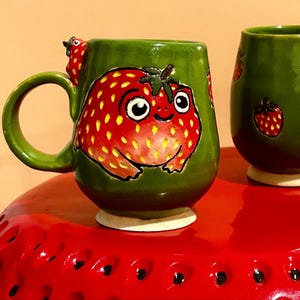 Könnte beinhalten: Eine grüne Keramiktasse mit einem skurrilen Erdbeerfrosch-Design. Die Tasse zeigt eine große rote Erdbeerfrosch-Illustration auf der Vorderseite, mit einer passenden Erdbeere am Griff. Die Tasse steht auf einer roten Oberfläche.