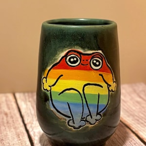 Può includere: Un bicchiere in ceramica verde scuro con un disegno di rana arcobaleno dipinto a mano. La rana è raffigurata in posizione seduta con un volto sorridente. Il bicchiere è cilindrico e sembra fatto a mano.
