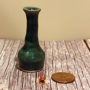 Puede incluir: Un pequeño jarrón de cerámica hecho a mano con un esmalte verde oscuro. El jarrón tiene un cuello estrecho y un cuerpo redondeado. El jarrón mide aproximadamente 5 cm de altura. Una moneda está al lado del jarrón para comparar el tamaño.