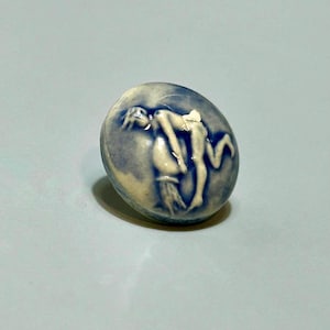 May include: A blue and white ceramic button with a relief sculpture of a figure. The button is oval-shaped and features a detailed depiction of a person in a dynamic pose. The color palette is primarily blue and white.