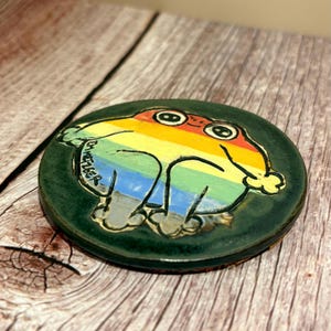 May include: A ceramic coaster featuring a colorful frog design. The frog is painted with rainbow colors and outlined in black, set against a dark green background. The coaster has a round shape and a glossy finish. The artist's signature is visible on the side.