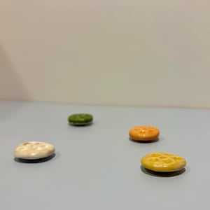 May include: Four small, ceramic, oval-shaped buttons in varying colours: off-white, green, orange, and yellow. Each button has a textured surface with small holes. The buttons are arranged on a light blue surface.