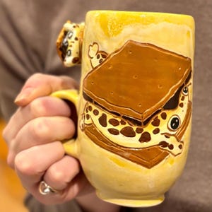 May include: A handmade ceramic mug with a yellow glaze. The mug features a detailed, hand-painted s'more design, with a graham cracker, marshmallow, and chocolate. The handle is also decorated with a s'more. A unique and fun kitchenware item.