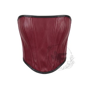 May include: A strapless corset top in a deep burgundy red color with a smooth, leather-like finish. The garment features vertical seam detailing throughout the front and is finished with a thin, dark black trim along the top and bottom edges.