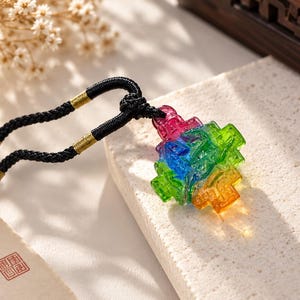 May include: A multi-colored glass pendant featuring a geometric, faceted design with vibrant red, green, blue, and orange hues. The pendant is suspended from a black braided cord accented with gold-toned metal wraps and small wooden beads.