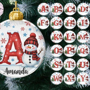 Puede incluir: Un adorno navideño personalizado con un muñeco de nieve y la letra "A" en rojo, con el nombre "Amanda" debajo. El adorno tiene una tapa dorada y está decorado con copos de nieve y un árbol de Navidad. Se muestran adornos adicionales con letras A-Z.