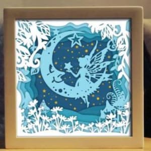 Op de afbeelding: Een ingelijst, gelaagd papierkunstwerk met een fee op een halve maan. Het kunstwerk gebruikt blauwe en witte tinten, in een gouden lijst. De fee houdt een ster vast, omringd door een sterrenhemel en bloemmotieven.