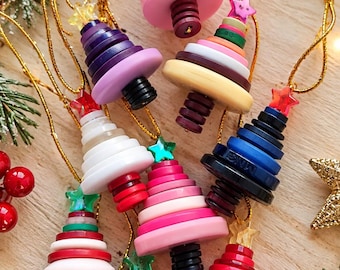 May include: A collection of Christmas tree ornaments crafted from stacked circular buttons in various colors including pink, red, white, blue, green, and purple. Each ornament is topped with a small plastic star and features a gold hanging cord.