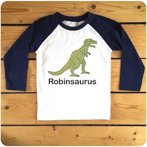 May include: White long-sleeve t-shirt with navy blue raglan sleeves. The shirt features a green dinosaur graphic and the text "Robinsaurus".