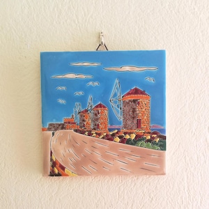 Può includere: Piastrella quadrata in ceramica con un'illustrazione di mulini a vento. L'immagine mostra mulini in pietra con tetti rossi, una strada e il mare sotto un cielo blu con nuvole e uccelli. Oggetto decorativo.