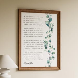 May include: A rectangular wall art print in a brown wooden frame featuring the poem The Road Not Taken by Robert Frost in black typewriter-style text, accompanied by a vertical watercolour illustration of green eucalyptus leaves on the right side.