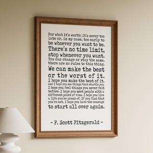 May include: A rectangular framed wall art piece featuring black typewriter-style text on a white background. The quote by F. Scott Fitzgerald begins with For what it's worth: it's never too late or, in my case, too early to be whoever you want to be.