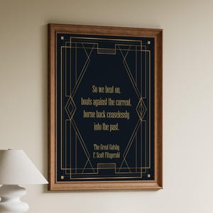Puede incluir: Un cuadro enmarcado con una cita de El Gran Gatsby de F. Scott Fitzgerald. El grabado tiene un fondo azul marino con diseños geométricos dorados de estilo art déco y texto. El marco de madera es de color marrón cálido.