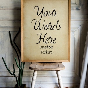 May include: A framed print with the text "Your Words Here Custom Print" on a light brown background. The frame is made of wood and has a light brown finish. The print is sitting on a wooden stool with white painted legs.