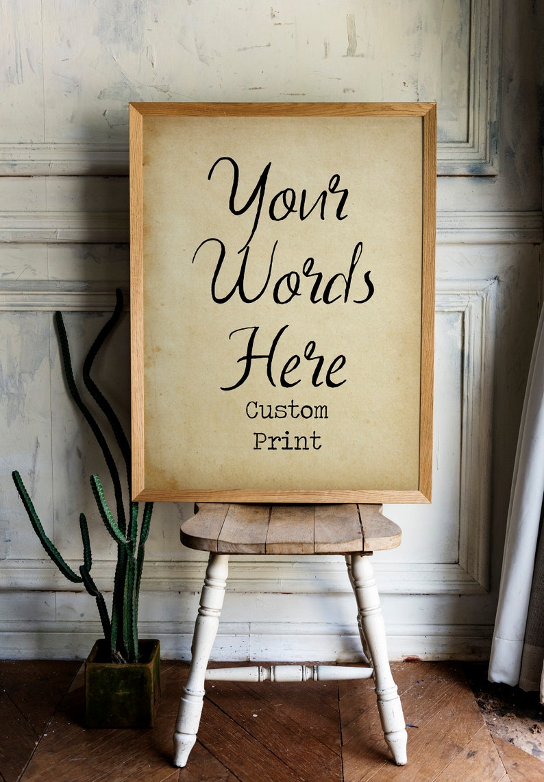 May include: A framed print with the text "Your Words Here Custom Print" on a light brown background. The frame is made of wood and has a light brown finish. The print is sitting on a wooden stool with white painted legs.