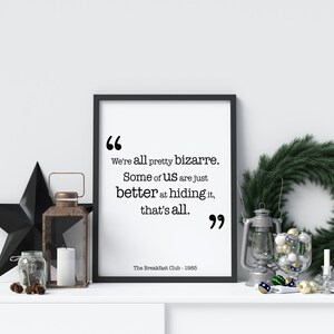 May include: Black and white printable quote from the film "The Breakfast Club" with the text "We're all pretty bizarre. Some of us are just better at hiding it, that's all." The quote is framed in a black frame and is sitting on a white surface with a black star, a glass jar with pine cones, a white candle, a lantern, and a wreath with baubles.
