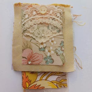 May include: A layered fabric scrap featuring a floral design, lace trim, and a white button. The fabric is in shades of yellow, orange, and brown.