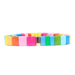 Op de afbeelding: Een kleurrijke armband gemaakt van vierkante kralen in tinten geel, oranje, roze, rood, groen, blauw en lichtblauw. De armband heeft een gouden accent in het midden.