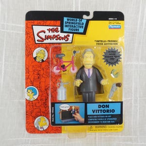 May include: A plastic action figure of Don Vittorio from The Simpsons, wearing a gray suit and a red tie. The figure is part of the World of Springfield Interactive Figure line and features Intelli-tronic voice activation. The packaging includes a small pink bicycle and a spray bottle.
