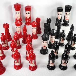 May include: A complete chess set featuring red and black pieces. The red pieces have a face design with a white base, while the black pieces have a similar design. The set includes rooks, pawns, knights, bishops, a queen, and a king.