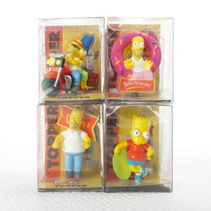 May include: Four clear plastic boxes containing yellow and red Simpsons character figurines. The boxes are labelled "Topper" and feature the characters Homer, Bart, Marge, and a donut.