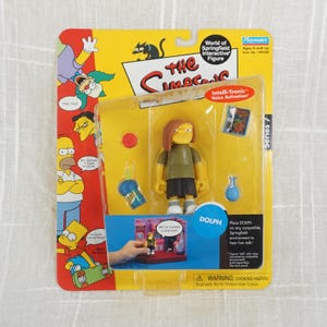 May include: A yellow and green action figure of Dolph from The Simpsons. The figure is holding a blue drink and has a yellow background with the text "Intelli-Tronic Voice Activation" and "Series 7".