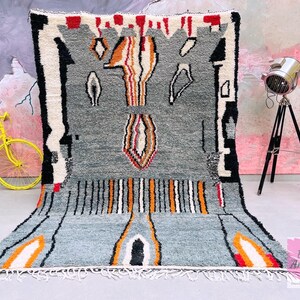 May include: A gray, white, orange, and red Moroccan rug with geometric patterns. The rug has a fringe border and is made of wool.