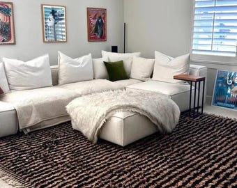 May include: A white sectional sofa with a fluffy white throw blanket and a small white ottoman. The sofa is in a living room with a brown and black striped rug and a small black side table.
