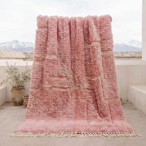 Pode incluir: Um tapete rosa grande e retangular com uma superfície texturizada e felpuda e franjas na borda inferior. O tapete é drapeado sobre uma estrutura, com vista para as montanhas ao fundo. O design do tapete apresenta padrões retangulares.