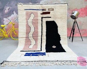 May include: A white, cream, pink, black, and blue abstract geometric patterned rug with a fringe border. The rug has a modern design with a wavy line, a black rectangle, and a blue rectangle.