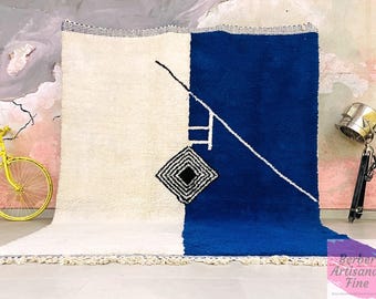 May include: A large, rectangular rug with a geometric design. The rug is divided into two sections: one is off-white, and the other is a vibrant blue. White lines and a black and white diamond shape add detail. The rug has a fringe along the bottom.