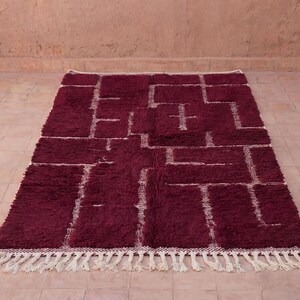May include: A rectangular burgundy rug with a geometric pattern in off-white. The rug has a thick, textured pile and a fringe along the bottom edge. The design features a series of connected rectangles and lines.
