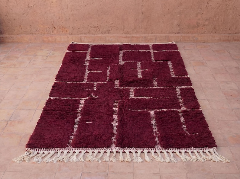 May include: A rectangular burgundy rug with a geometric pattern in off-white. The rug has a thick, textured pile and a fringe along the bottom edge. The design features a series of connected rectangles and lines.