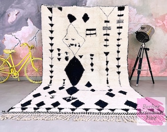 May include: Black and white Moroccan rug with a geometric pattern. The rug has a fringe border and is made of wool. The rug is on a gray floor in front of a pink and gray wall.