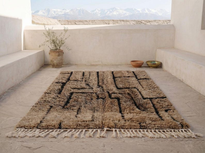 May include: A rectangular, beige rug with a geometric black design and fringe. The rug is placed on a concrete patio, with a view of mountains in the background. A potted plant and bowls are also visible.