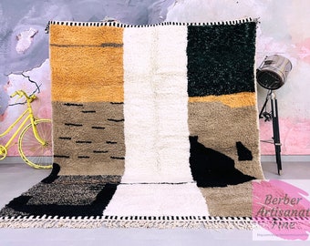 May include: A hand-woven wool rug with a geometric pattern in shades of beige, black, white, and mustard yellow. The rug has a fringe border.