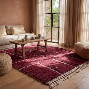 May include: A living room scene featuring a burgundy geometric rug with a fringed edge, a wooden coffee table, and a beige sofa with decorative pillows. Two woven poufs and large windows complete the cozy space.