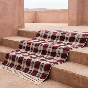 Puede incluir: Una alfombra rectangular con un patr&oacute;n de cuadros en tonos burdeos, crema y gris, con flecos en un borde. La alfombra est&aacute; colocada sobre unos escalones de hormig&oacute;n, creando una est&eacute;tica texturizada.