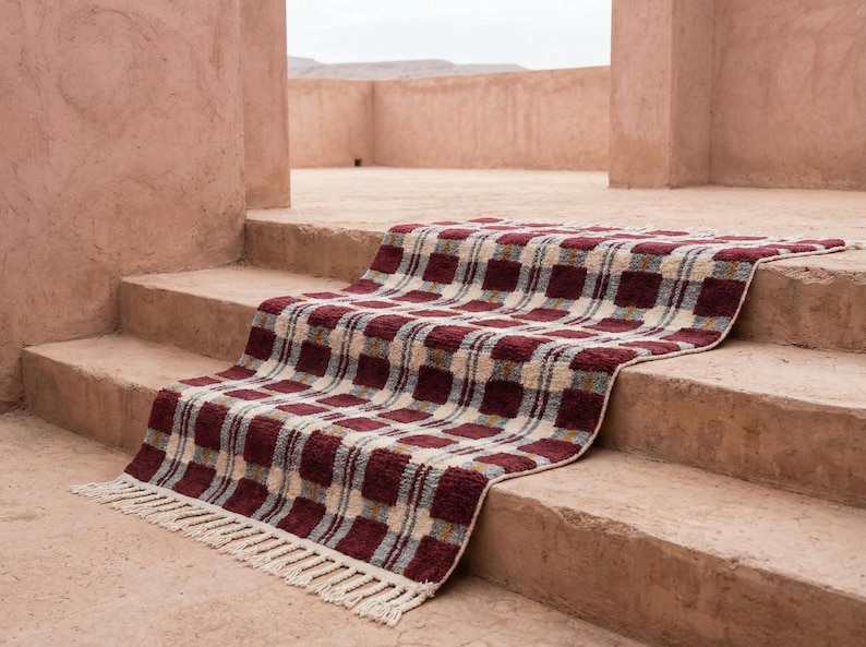 Puede incluir: Una alfombra rectangular con un patr&oacute;n de cuadros en tonos burdeos, crema y gris, con flecos en un borde. La alfombra est&aacute; colocada sobre unos escalones de hormig&oacute;n, creando una est&eacute;tica texturizada.