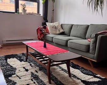 May include: A living room with a gray sofa, a wooden coffee table with a red runner, and a black and white rug. The coffee table has a wooden frame and a dark brown top.
